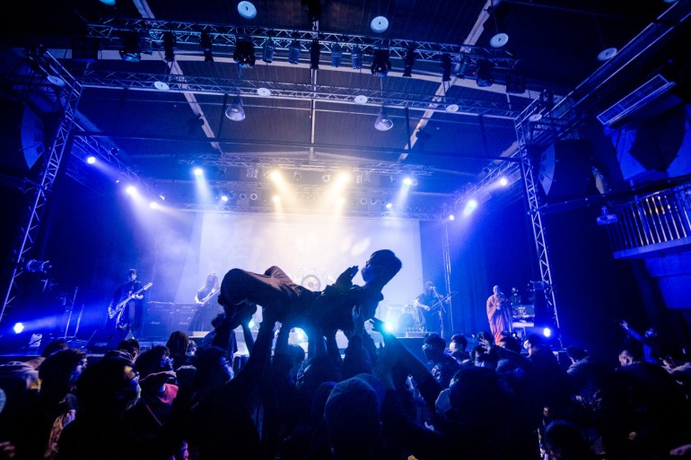 A fan crowd surfing at a Dharma gig. The fan is in the lotus position and the crowd is holding him up. The band are playing on stage. Everything is bathed in blue light