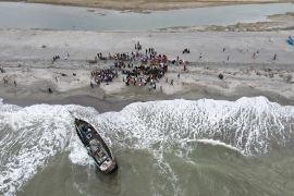 An aerial view of a boat amid waves crashing on the beach and dozens of passengers ashore.