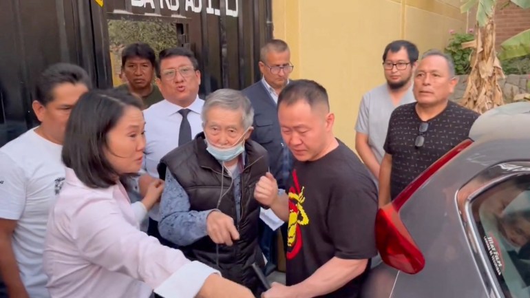 Alberto Fujimori, an elderly man wearing a black puffer vest and a face mask, is greeted outside the prison gates by his daughter Keiko and son Kenji. Other officials look on. Keiko points, presumably to guide her father to a waiting vehicle visible in the corner of the photo.