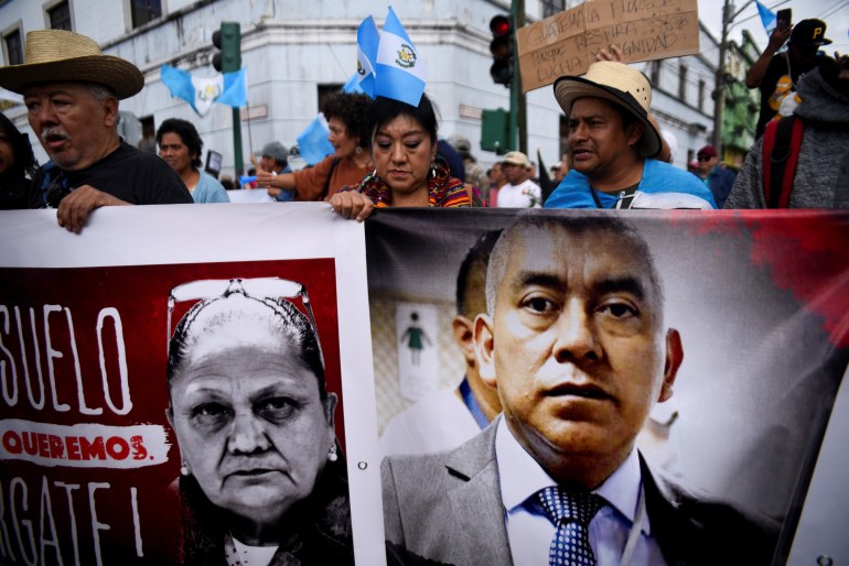 Protesters in Guatemala hold up banners with the Attorney General Maria Porras's face and that of her prosecutor Rafael Currichiche. Guatemalan flags wave behind them.