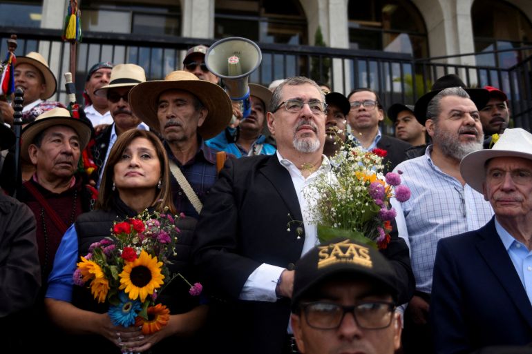 Bernardo Arevalo and Karin Herrera each hold a bouquet of flowers as part of a protest. They stand in front of a government building with white columns, surrounded by a cast-iron fence.