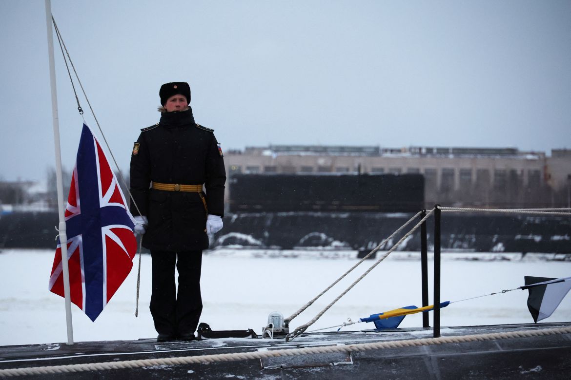 A sailor attends a flag-raising ceremony on the nuclear-powered submarine Emperor Alexander III. There is a flag flying next to him. There is snow on the ground.