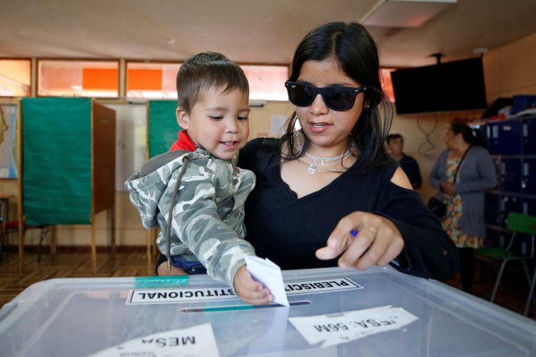 A woman in sunglasses holds a baby up to a ballot box, where the little child slips a ballot into the slot.