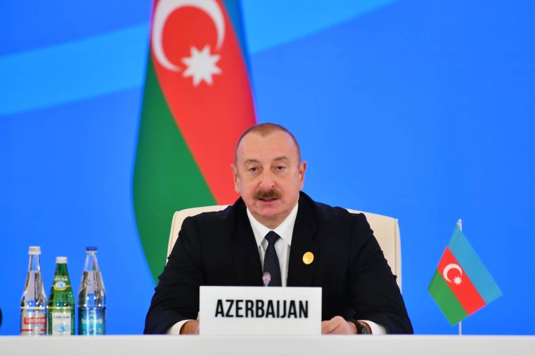 A photo of Ilham Aliyev sitting at a table with a sign that says "Azerbaijan" in front of him with three bottles of water next to him as well as a small Azerbaijan flag next to him and a bigger Azerbaijan flag behind him.