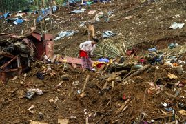 A woman picking through the debris after a military attack on a displaced persons camp in Myanmar. Things are strewn across the muddy hillside.