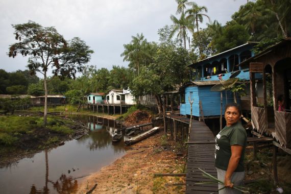 A woman stands on a walkway made of wooden planks that runs along a dried-up river. Houses on stilts sit along the river bank.
