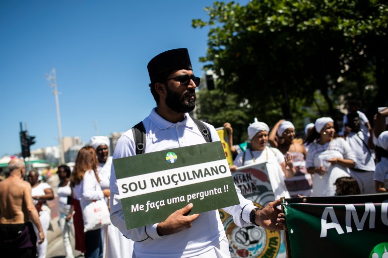 A man walks in a demonstration, carrying a sign that reads, "Sou Muçulmano. Me faça uma pergunta!"