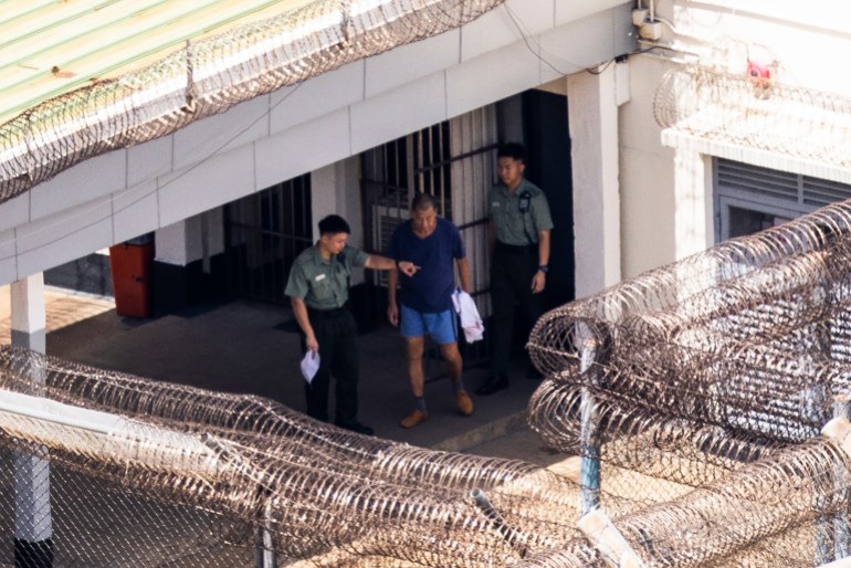 Jimmy Lai in blue prison uniform seen in the yard of the Stanley jail. It is surrounded by high fences topped with razor wire. He is accompanied by two prison officers