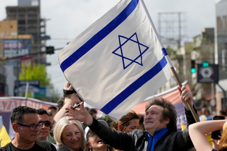 Javier Milei, standing outdoors amid a crowd, holds up an Israeli flag.