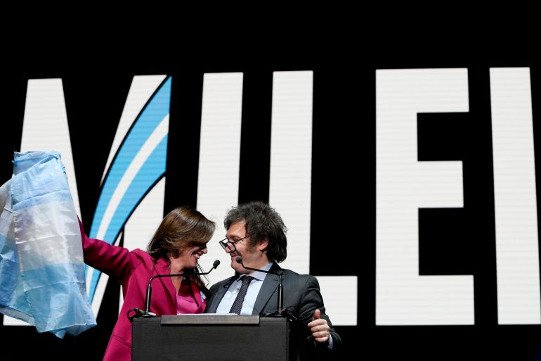 Javier Milei in a dark suit and Victoria Villarruel in a red suit stand in front of a screen with giant, capitalized letters that read, "Milei." Villarruel holds up an Argentine flag.
