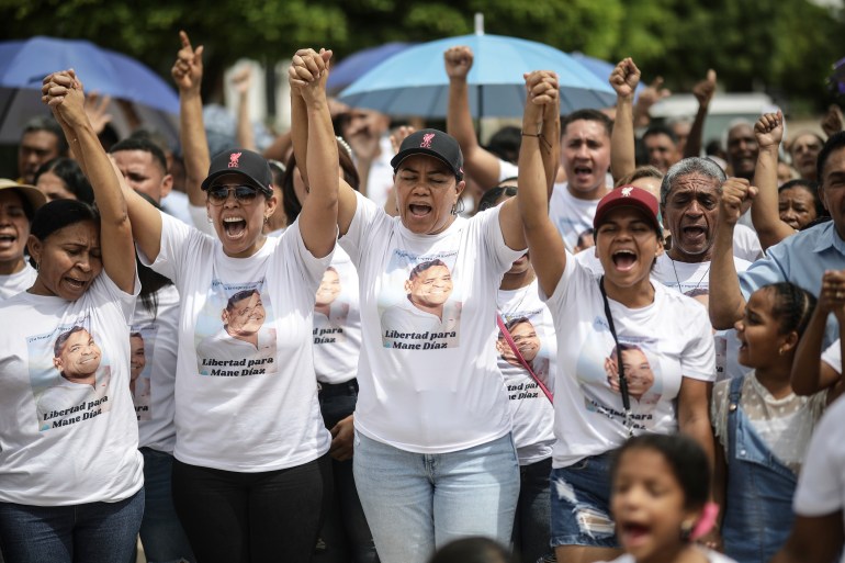 Women in ball caps and T-shifts lock hands and lift their arms in the air, as they protest to push for the release of Luis Diaz's father. The father's face is printed on each of their T-shirts.