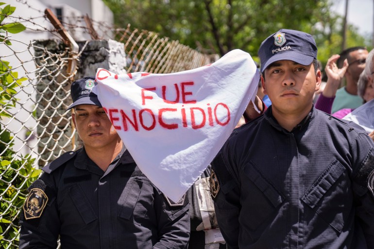 Two police officers — dressed in black uniforms with gold crests on the arm and black caps on their head — stand side by side as someone behind them holds up a white bandana that reads: "Fue Genocido"