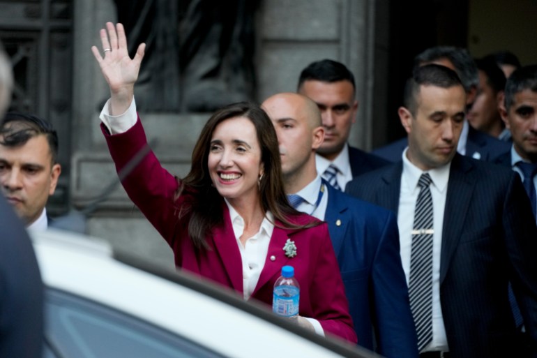 Victoria Villarruel, holding a water bottle and wearing a red suit, waves with one arm stretched high in the air as she prepares to enter a waiting vehicle.