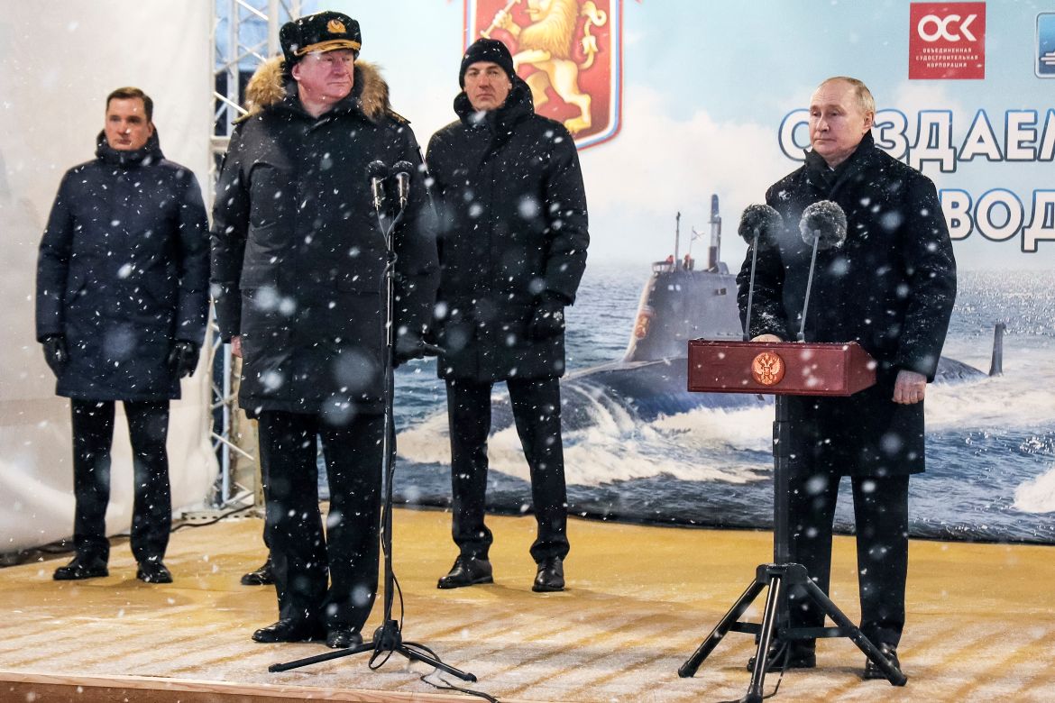 Putin speaking at an outdoor stage. Admiral Nikolai Yevmenov, Commander-in-Chief of the Russian Navy, is to his right. Other senior naval officers are also on stage. It is snowing.