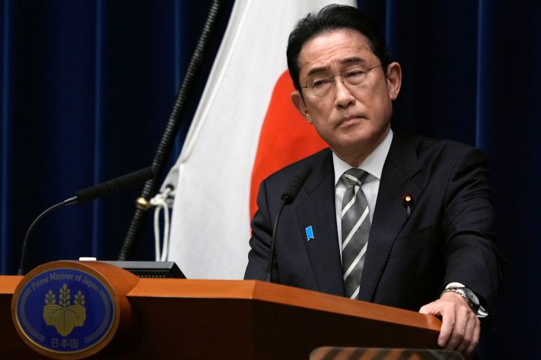 Japanese Prime Minister Fumio Kishida. He is standing at a lectern in front of a Japanese flag. He looks serious.
