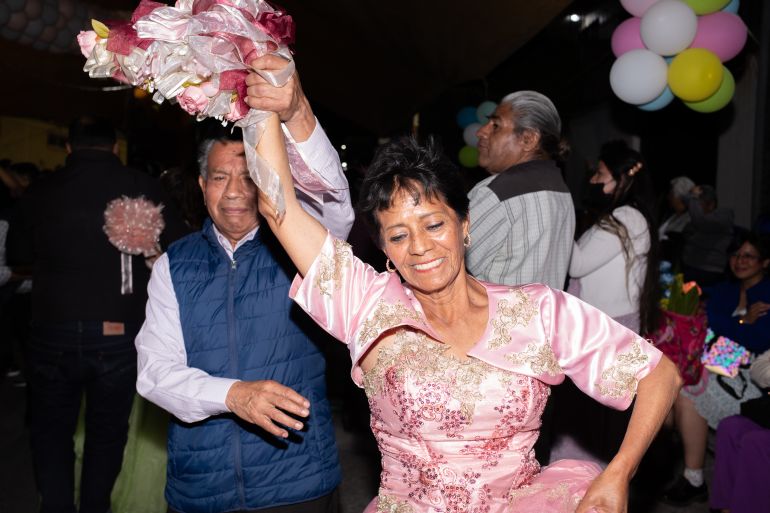 A woman in a pink dress is twirled by her dance partner, one arm lifted into the air as she perches the other on her hip.