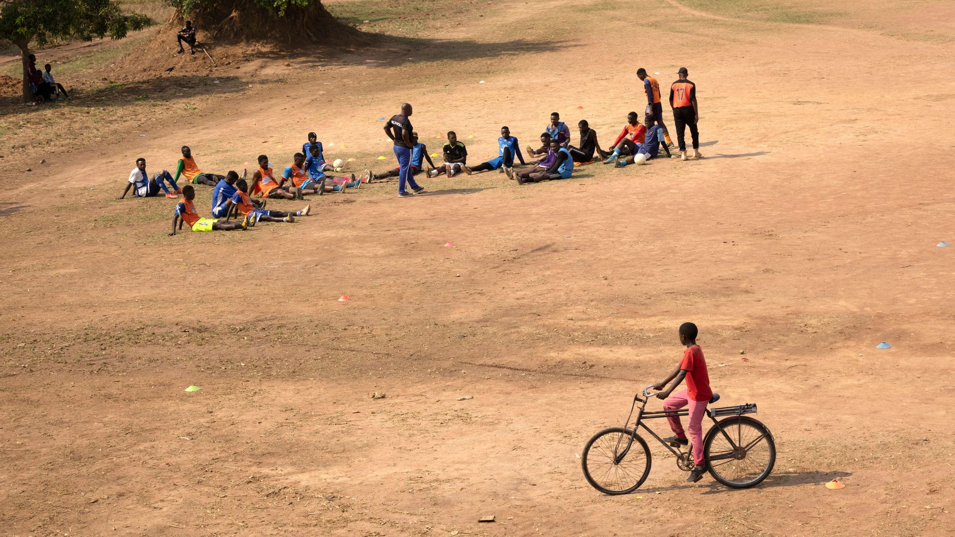 A child on a bicycle passes by as a football team gather for training on a field in a refugee camp in Zambia