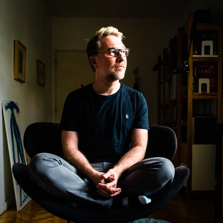 A man in glasses, a black T-shirt and jeans sits on a table, looking toward the light.