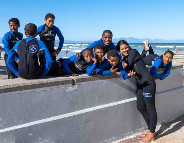Kids in wetsuits pose on the wall with their instructor