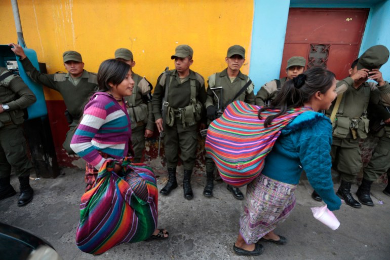 Women in traditional textiles — with rainbow-colored stripes — walk in front of a line of military members on a Guatemalan street.