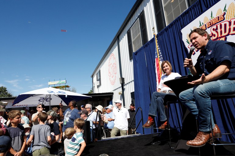Kim Reynolds and Ron DeSantis sit on a blue-draped stage with an American flag and a corn-themed logo advertising "Kim Reynolds's fair-side chats." People mill around below the stage, as the Iowa State Fair continues around them.