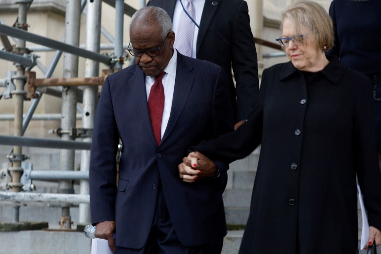 Clarence Thomas and Ginni Thomas, dressed in black for a funeral, hold hands as they walk down cathedral steps.