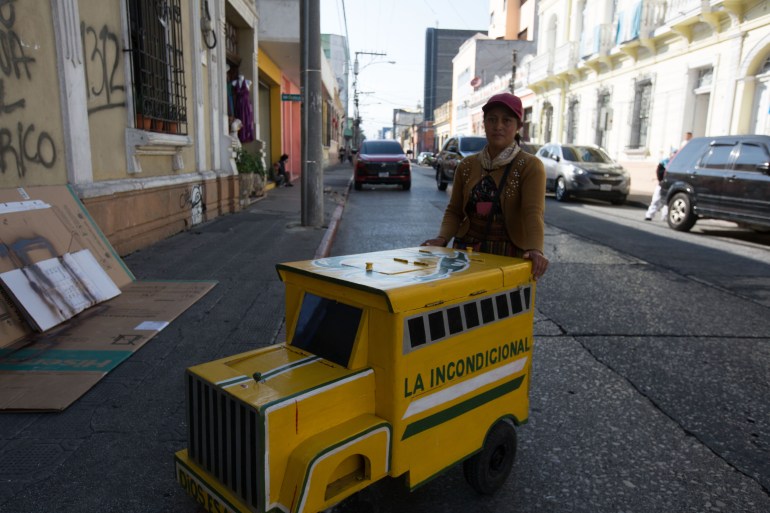 A woman stands behind a small, mobile freezer cart in the shape of a yellow school bus in the streets of Guatemala City. The school-bus cart has an opening at the top, where the woman can reach in to fetch ice cream for customers.