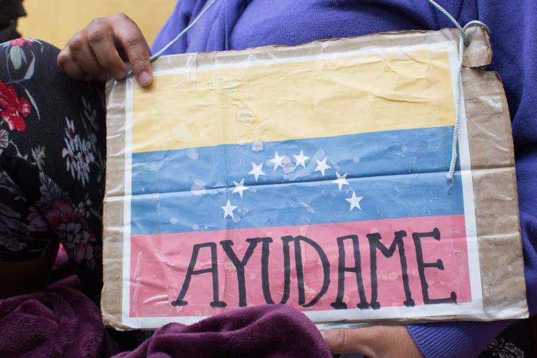 A Venezuelan migrant, whose face is not visible in the photograph, holds up a sign that shows the Venezuelan flag with the phrase, "Ayudame" or "Help me."