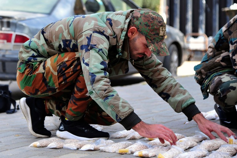 A Syrian soldier kneels over rows of captagon in little packets, arranged on the pavement.