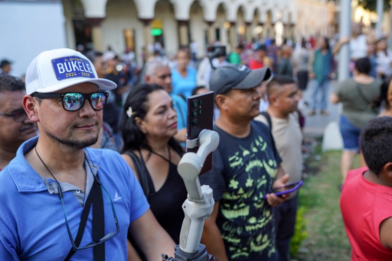 A man wearing a blue polo shirt and a baseball cap emblazoned with "Bukele 2024-2019" holds up a selfie stick in a crowd on the street.
