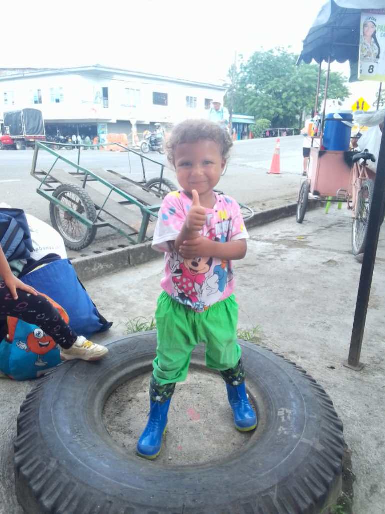 Emiliannys stands in the center of a tire that lies flat on the ground. She wears blue rain boots, green pants and a pink shirt.