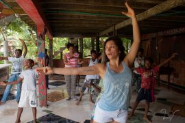A woman in a blue tank top printed with the name of Miami, Florida, lifts one arm above her head and holds the other outstretched by her side, as she practices salsa moves for a class of students who mimic her.