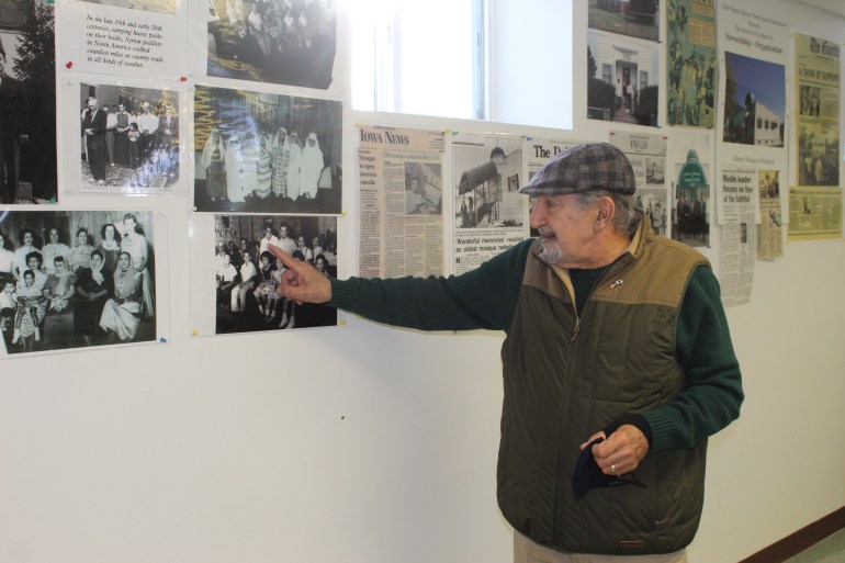 A man in a plaid flat cap and a cold-weather vest points to an image of himself on a wall lined with newspaper clippings and old photos.