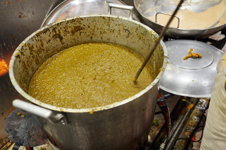 A large pot of saag simmers as a volunteer uses a large drill to stir the dish thoroughly [Sonny Thakur/Al Jazeera]