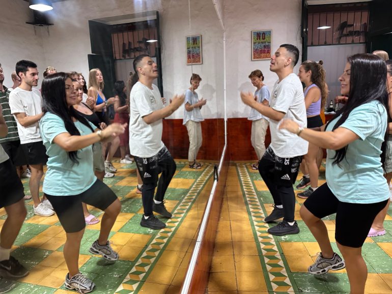 Two dance instructors stand at the front of a classroom in front of a mirror the size of a wall. As they dance, students behind them mimic their steps.