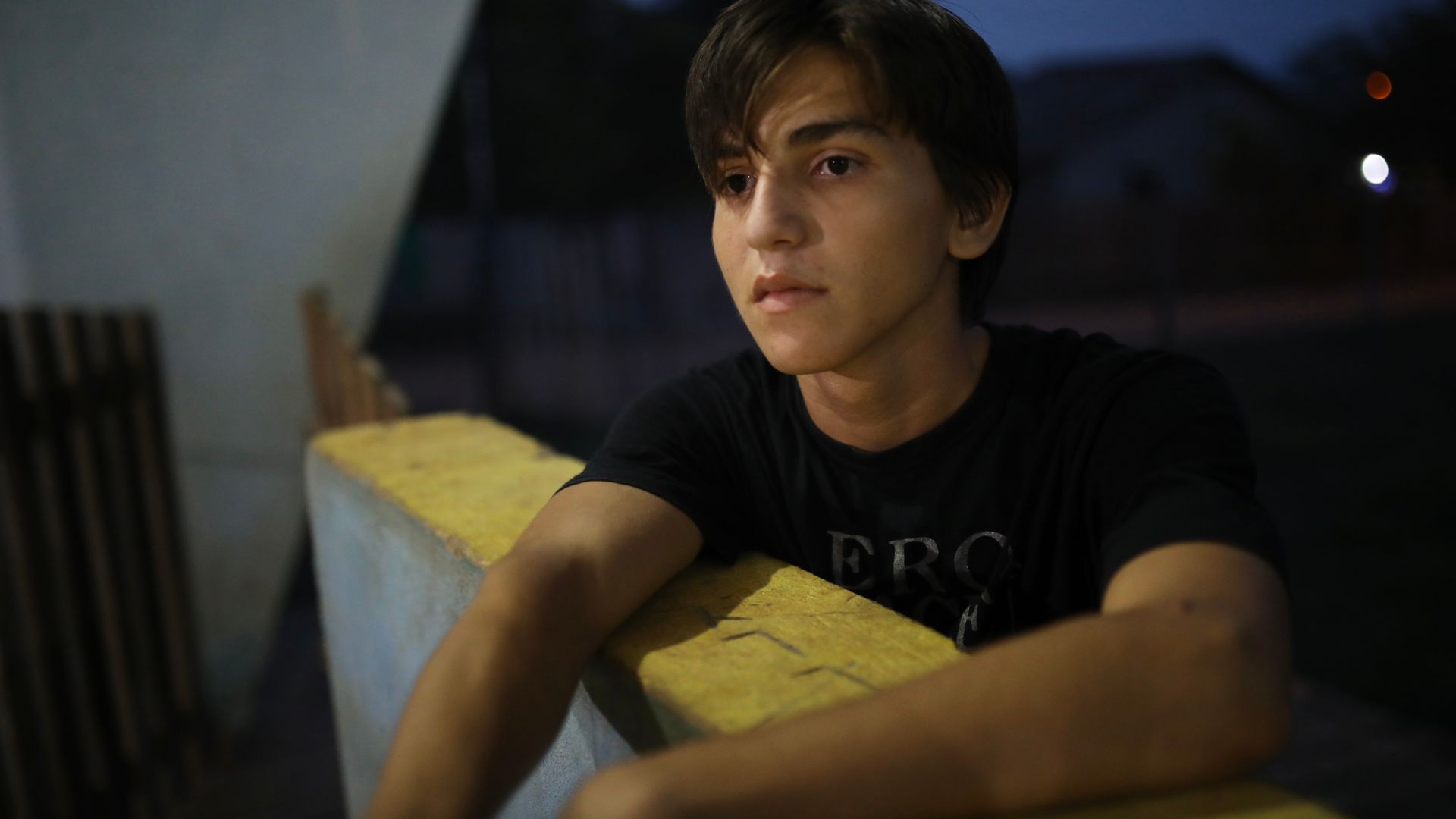A teenage boy leans against a concrete wall. Behind him is a dark-blue nighttime sky.
