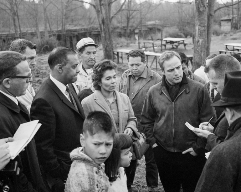 A crowd gathers in an outdoor space in Washington state, with trees and a picnic table visible in the background. Marlon Brando stands to one side, speaking to a reporter who takes notes on a notepad. Next to him is Indigenous leader Janet McCloud, and in the foreground are a pair of children.