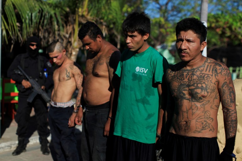 A line of young men — some shirtless with tattoos, another with a green shirt — stand with bowed heads outside in Puerto el Triunfo.