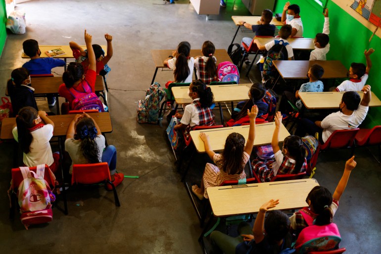 Seen from above, students sit in a classroom, two per desk table, and raise their hands enthusiastically.