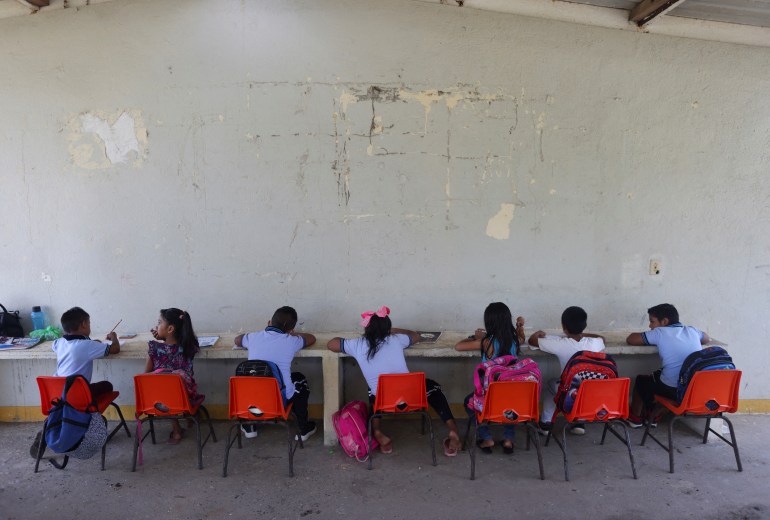 A row of young pupils sit in orange plastic chairs at a long desk placed against a peeling wall in a Mexico school.