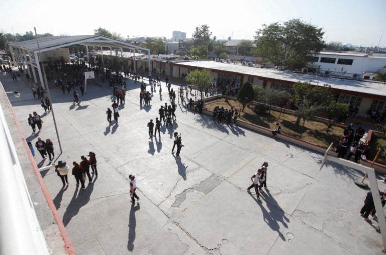 Students are seen from above walking in a paved courtyard between two school buildings. A basketball hoop is seen to one side.