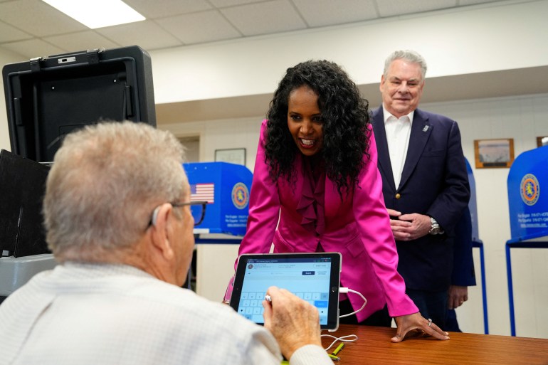 Mazi Melesa Pilip, in a bright pink blazer, leans over a table and smiles as a poll worker on the other side looks at an electronic tablet.