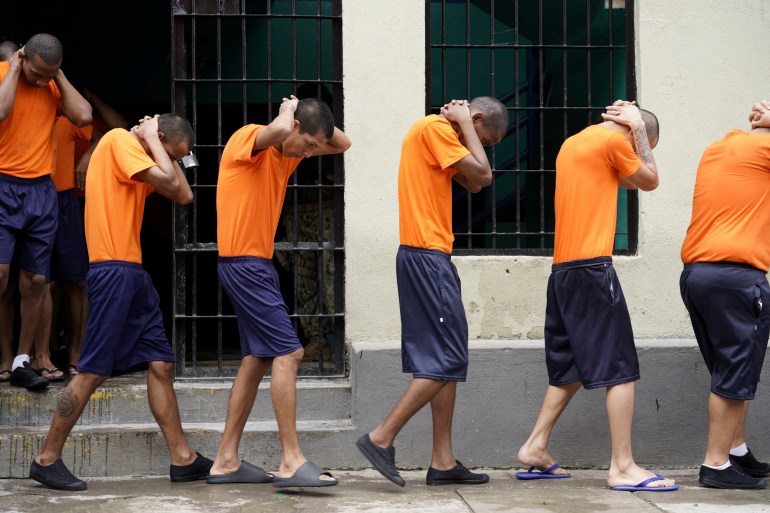 Prisoners in orange shirts and shorts walk in a single-file line, their hands behind their downward-facing heads.