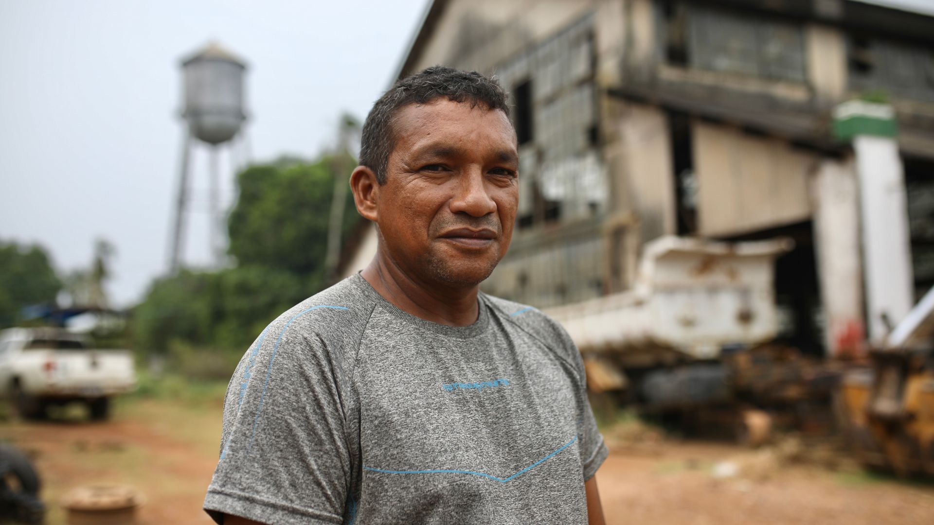 A man in a grey T-shirt stands in front of the abandoned warehouses at Fordlandia, an industrial town in the Amazon. A water tower is also visible behind him.