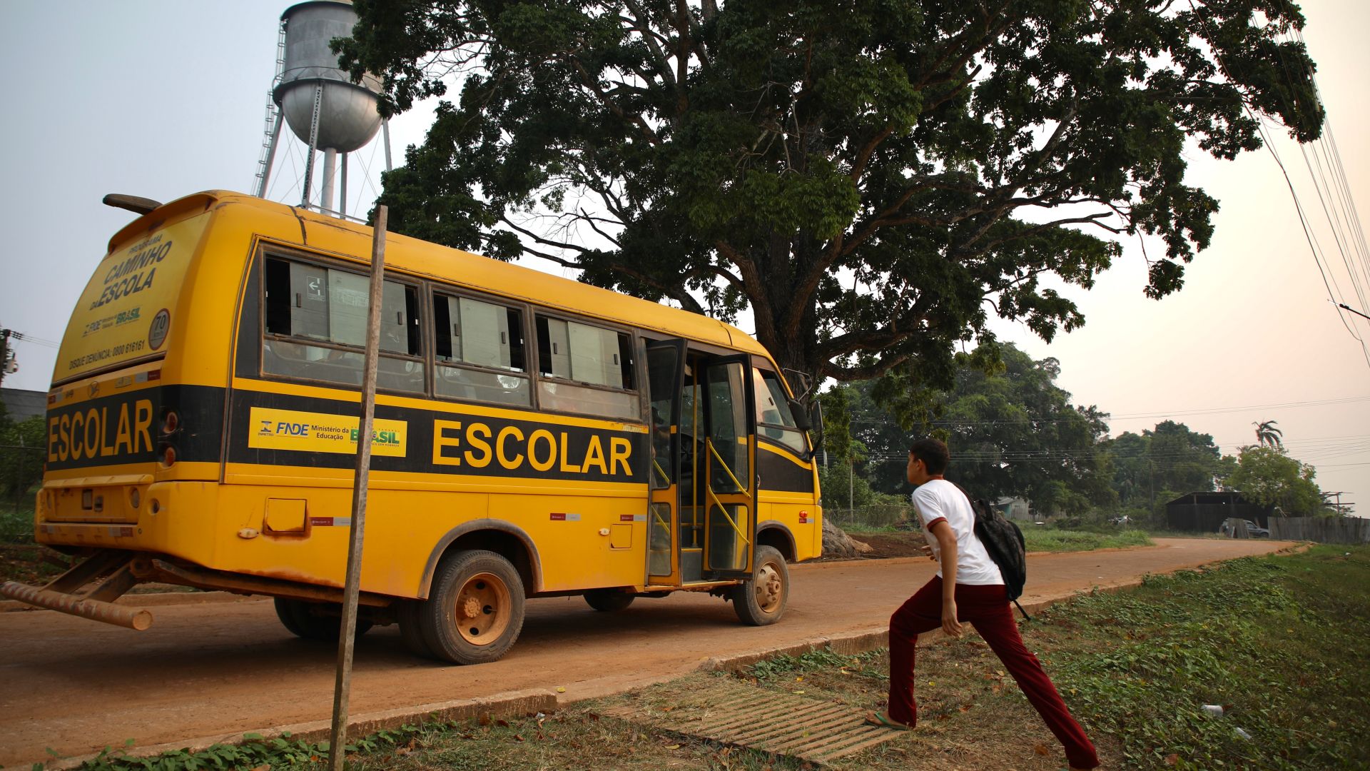 A young student takes wide strides to reach an orange school bus waiting for him on a dirt road.