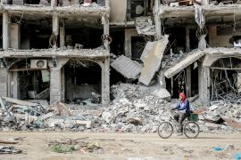 A Palestinian man rides a bike past a destroyed building [Omar Qattaa/Anadolu]