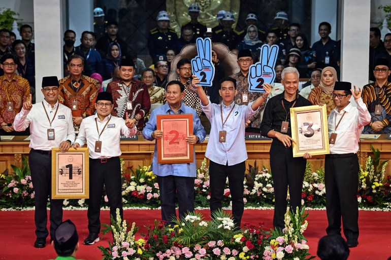 The presidential candidates with their running mates. They are holding up placards with their candidate numbers - 1, 2 and 3. Gibran is holding up two pale blue hands with the number two written on them.