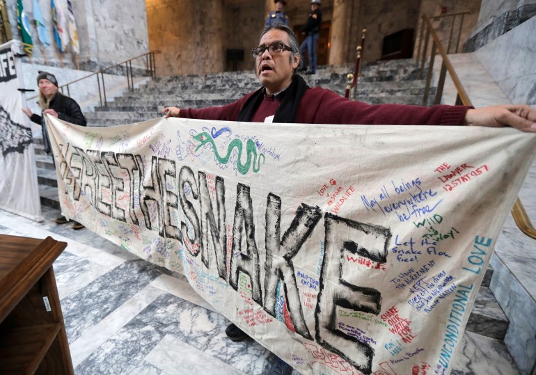 An Indigenous activist in the state house of Washington State holds up a giant handwritten banner that reads, "Free the Snake," in reference to the Snake River dams