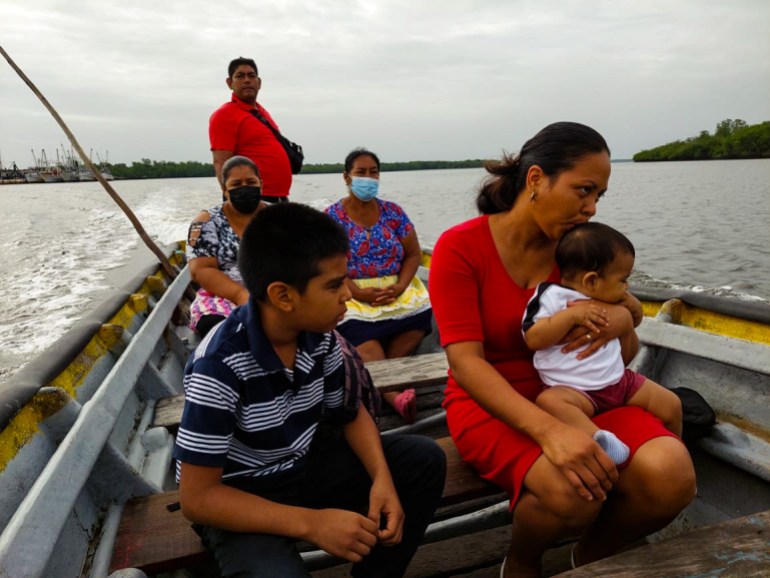 A man in a red shirt stands at the back of a small boat as it crosses the water. In front of him are two older women, a child, and a woman with a baby on her lap.