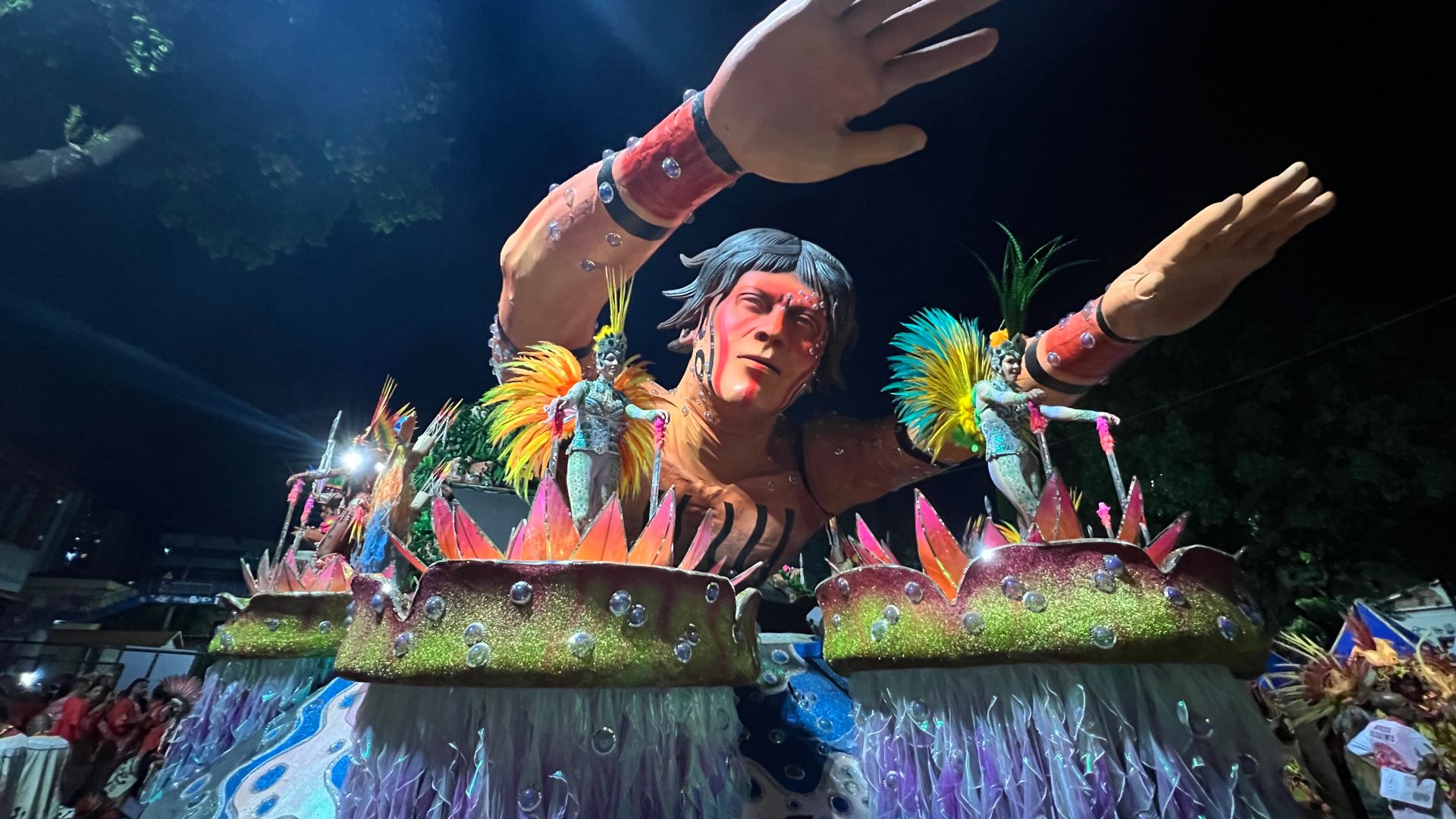 A float at the Sambadrome in Rio de Janeiro depicts a Yanomami person with his arms outstretched. On either side of him are platforms where dancers perform, dressed in blue sequins in a riot of yellow feathers emerging like wings from their backs.